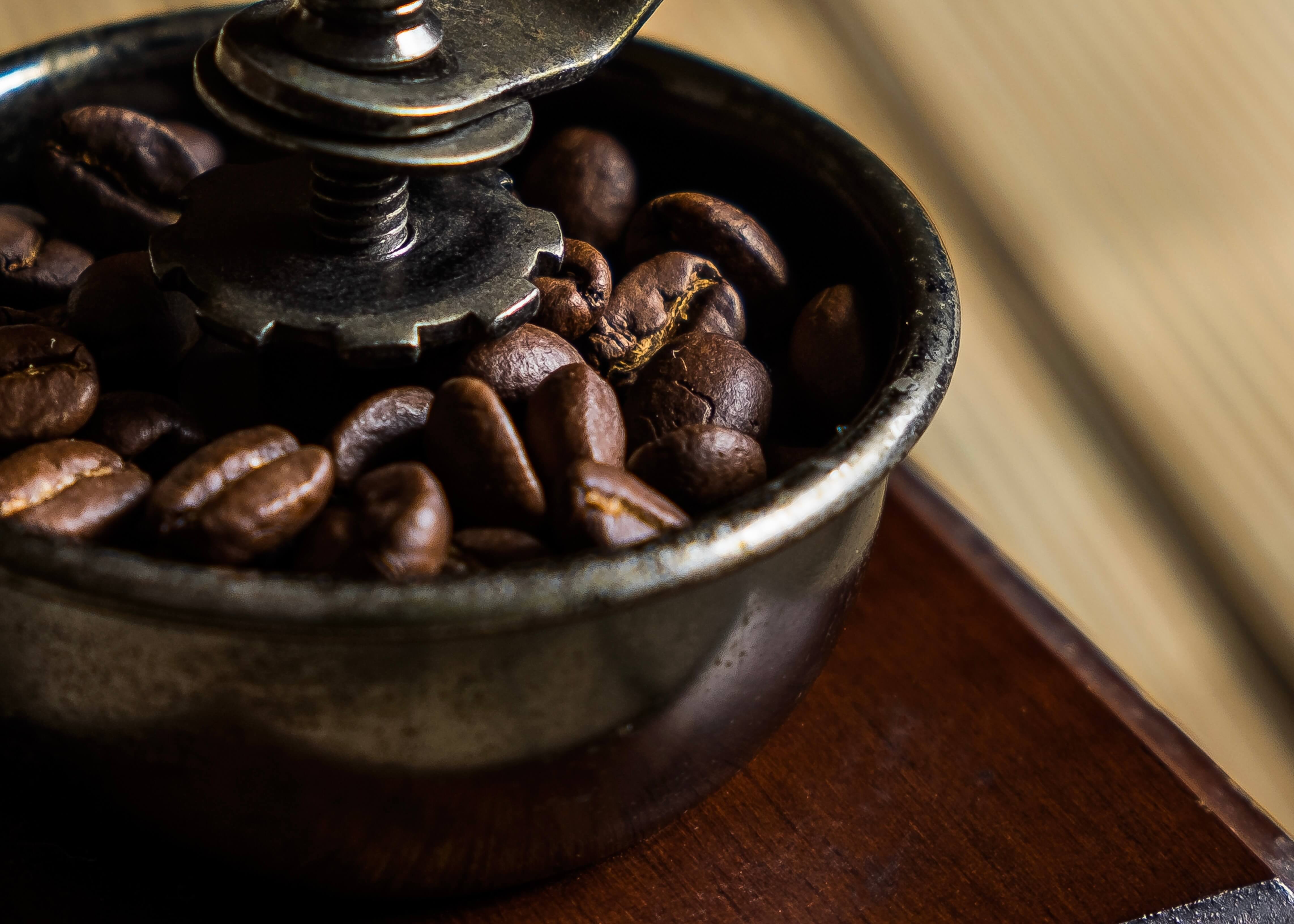 Nærbillede af ristede kaffebønner i en metalkværn, hvor en del af mekanismen er synlig ovenover. Den let slørede baggrund fremhæver teksturen - ideel til plakaten "En kaffekværn i drift" af Posters, som hylder kaffehåndværket.