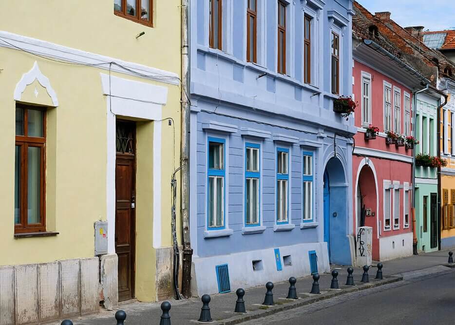 Plakaten "Farvede huse i række" fra Posters viser en livlig gade med gule, blå og lyserøde bygninger med blomsterdekorerede vinduer og pæne sorte pullerter langs fortovet.