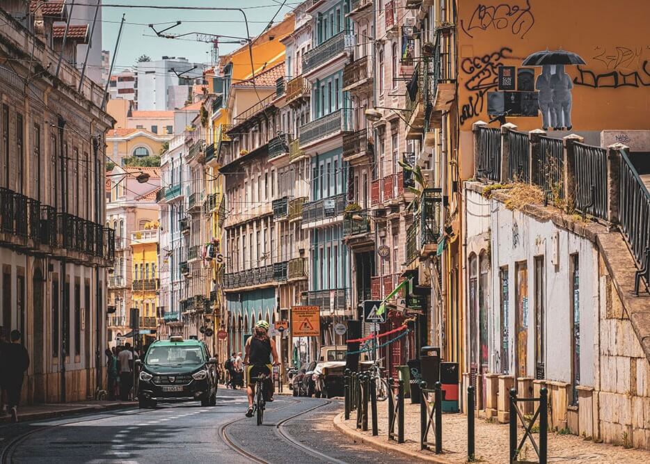 Farvefuld Gade by Posters viser en livlig europæisk gade med gamle bygninger, balkoner og plakatbeklædte vægge. En cyklist kører foran, mens biler, fodgængere og gadeskilte fylder den farverige scene under en overskyet himmel.