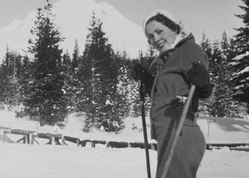 En smilende kvinde i vintertøj poserer med skistave på en sneklædt skråning blandt snedækkede træer og bjerge. "Golden Rose Ski Classic Princess" fra Posters indfanger den tidløse charme ved en klassisk skiferiescene.