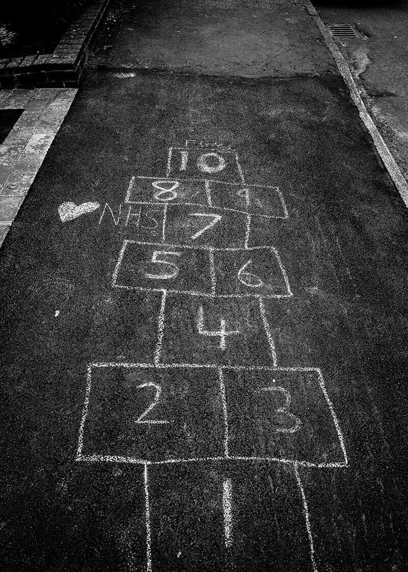 Hopscotch Game in Monotone" fra Posters viser et hvidt kridtnet nummereret 1-10 på fortovet med et hjerte og et NHS ved siden af. Dette sort-hvide print fremkalder nostalgi - perfekt som en minimalistisk plakat til moderne indretning.