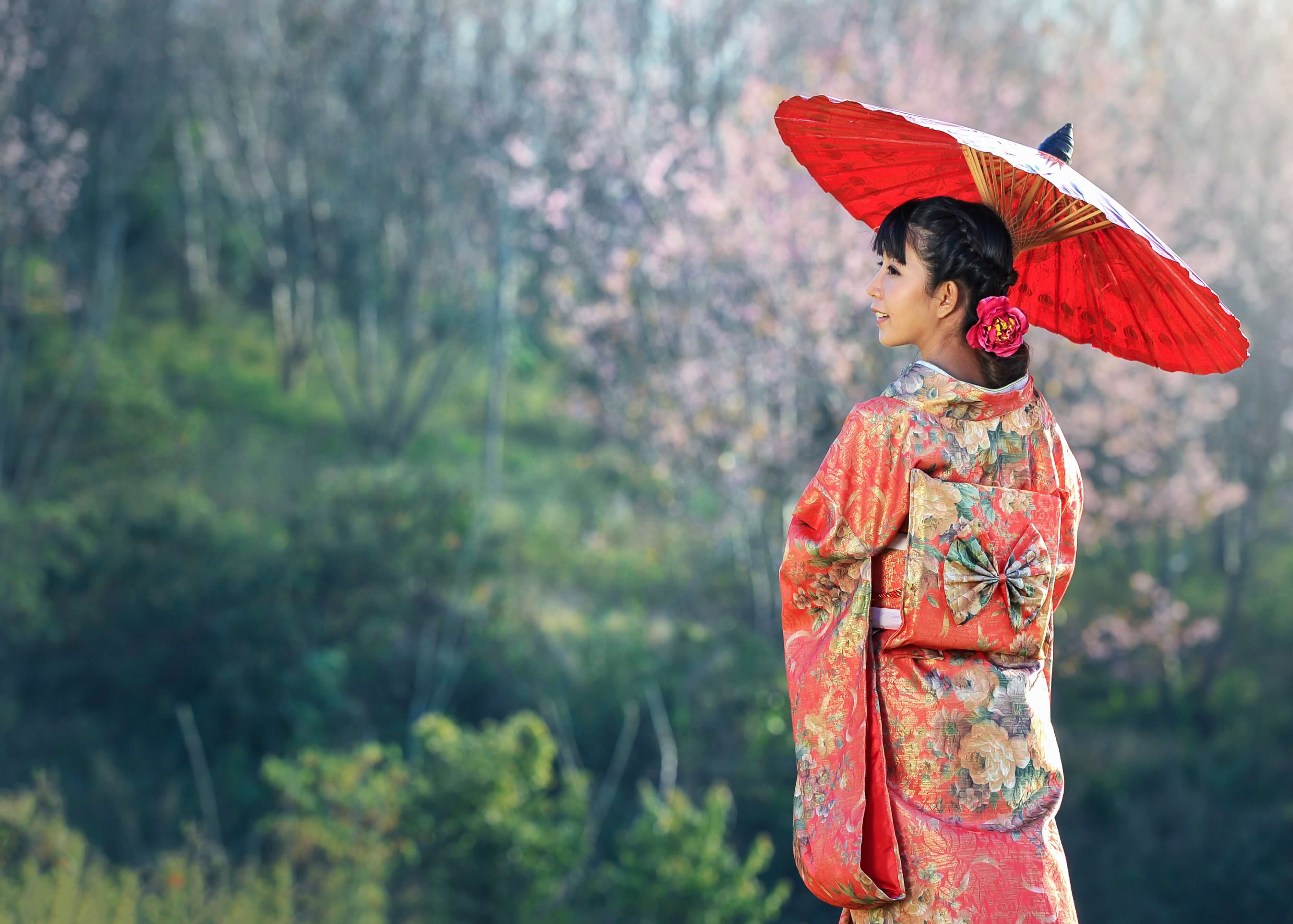 Japansk kvinde nyder udsigten"-plakaten fra Posters viser en smilende kvinde i farverig kimono med rød parasol omgivet af blomstrende træer og grønt - et billede med harmonisk atmosfære.