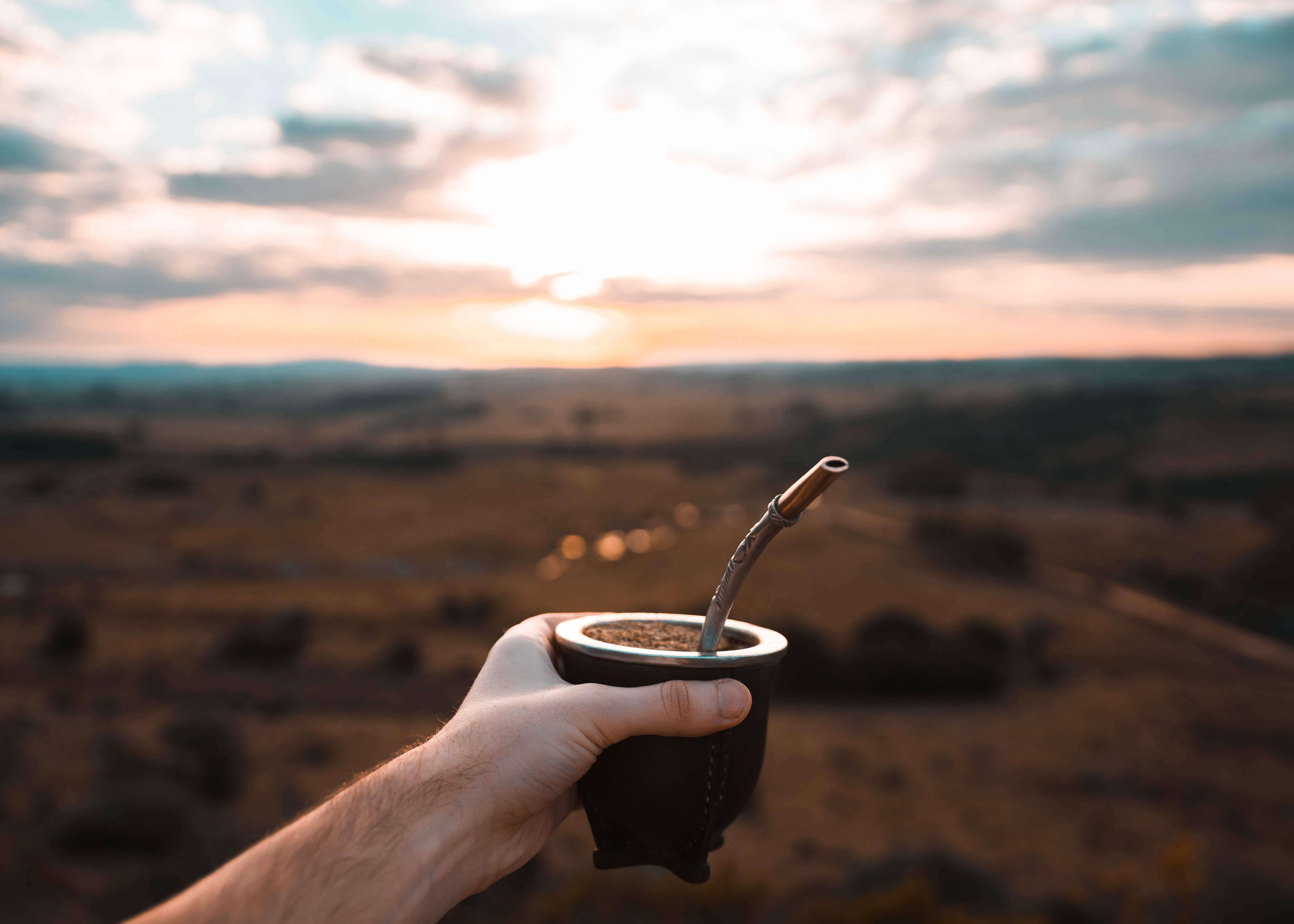 En hånd holder Kaffeskål by Posters - en traditionel mate-kalebasse med metalstrå - hævet over græsklædte sletter og en levende, skyfyldt solnedgang som en scene fra en rejseplakat.