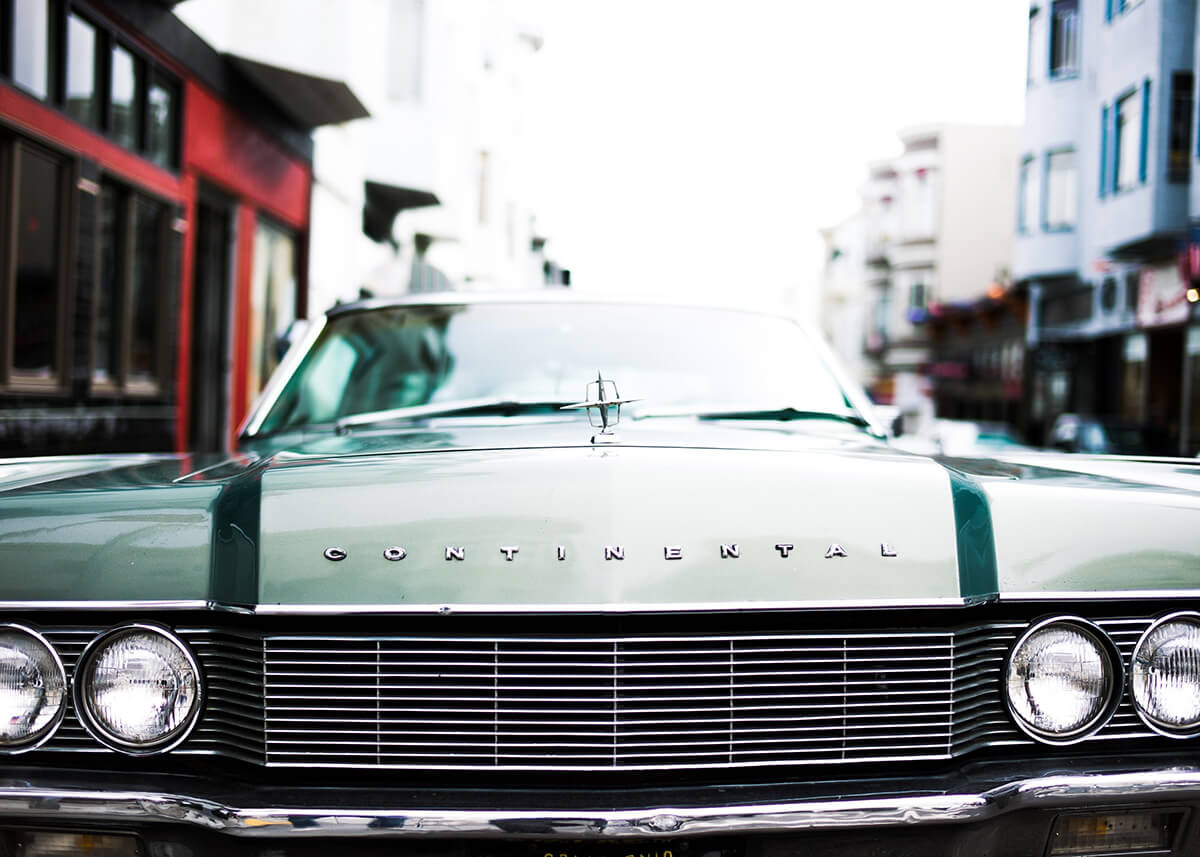 Nærbillede af en vintage grøn Lincoln Continental parkeret på en gade i byen, ideel til en Lincoln Continental plakat eller retro bilplakat. Bilens kølergrill, forlygter og Continental-badge skiller sig ud fra slørede bygninger.