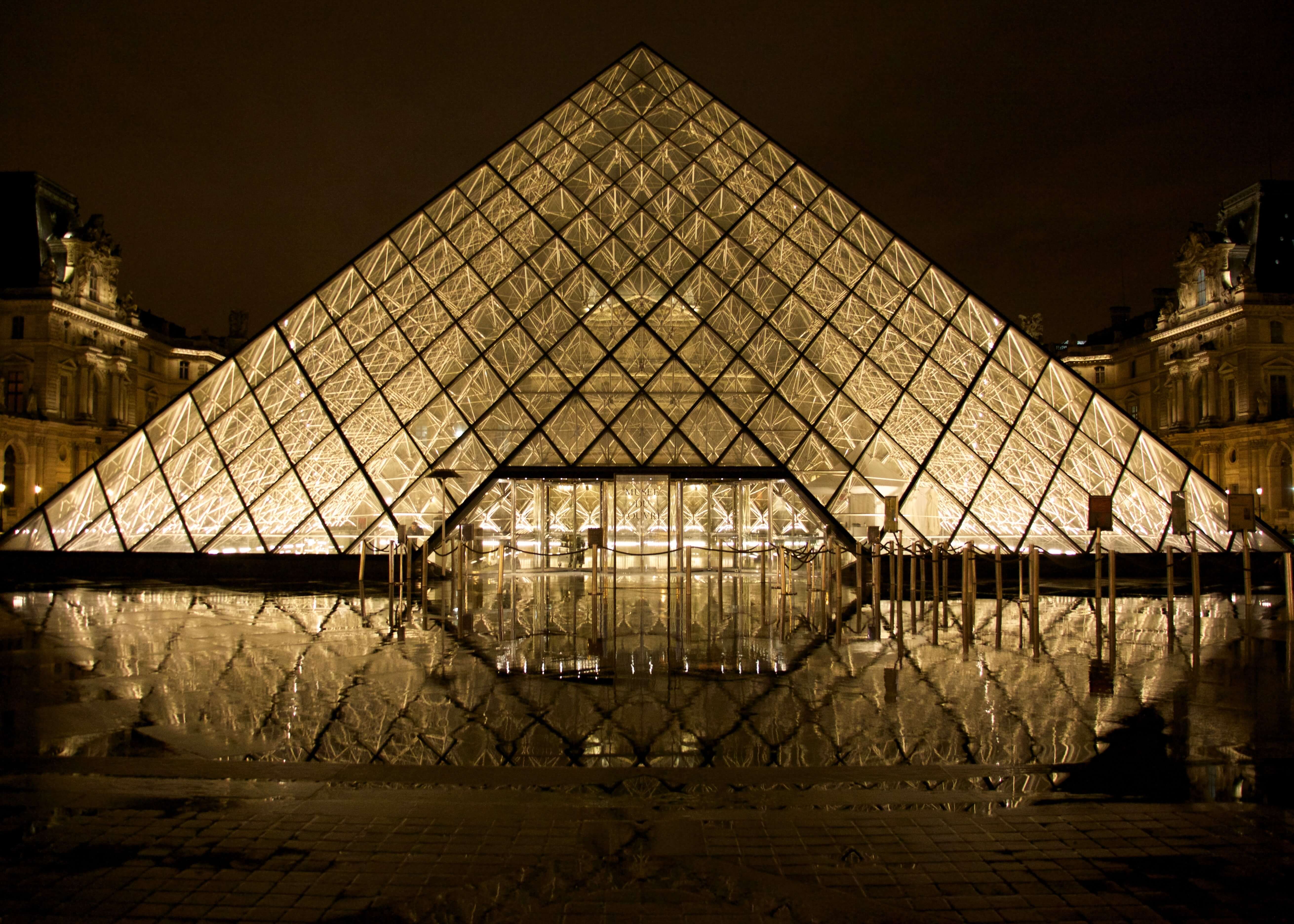 Louvre Glass Pyramid by Posters viser det oplyste vartegn i Paris om natten, hvor dets refleksioner skinner i vandet nedenfor mod en mørk himmel og indfanger det dramatiske udseende af en slående plakat.