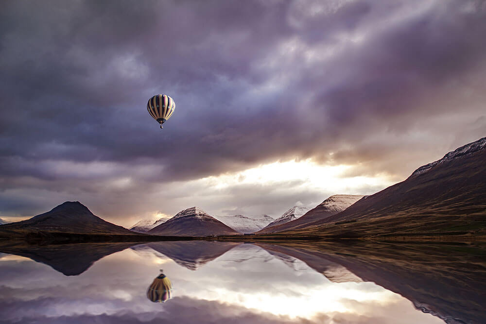 Plakaten "Luftballon over bjerge i lilla" fra Posters viser en stribet varmluftsballon, der svæver over en rolig sø med sneklædte bjerge under en dramatisk himmel, hvis refleksioner spejler sig i det stille vand nedenunder.