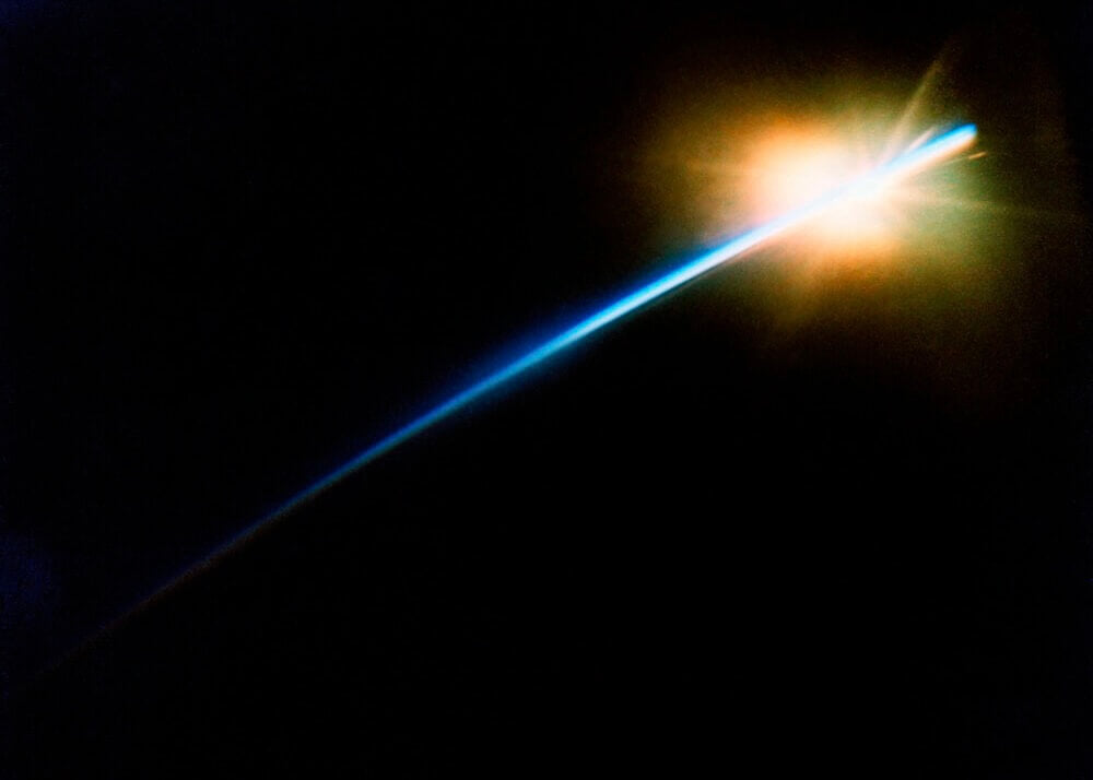 En strålende blå og orange stribe bryder frem diagonalt over en sort baggrund på plakaten "Lysstråle i Mørket" fra Posters, der ligner en komets glød i mørket.