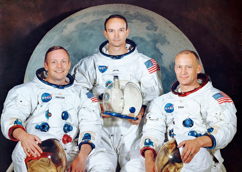 Tre astronauter i NASA-rumdragter står foran en stor "Månelandingen"-måneplakat fra Posters med hjelme og udstyr. På deres ærmer er der lapper med det amerikanske flag, mens de kigger ind i kameraet.