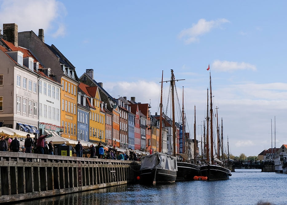 Nyhavn en forårsdag by Posters indfanger den livlige havnefront i København med farverige bygninger, fortøjede sejlbåde og menneskemængder langs kajen under en delvis overskyet himmel - perfekt dansk charme til din væg.