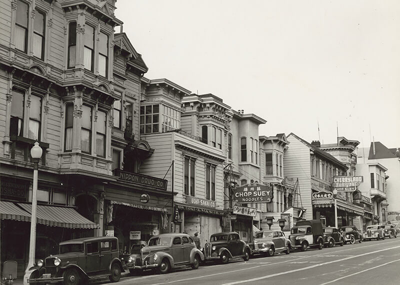 Sort-hvidt foto af en travl gade i San Francisco med veteranbiler og butiksfacader. "San Francisco, California" af Posters - ideelt for elskere af vintage bybilleder og FSC-certificerede print.