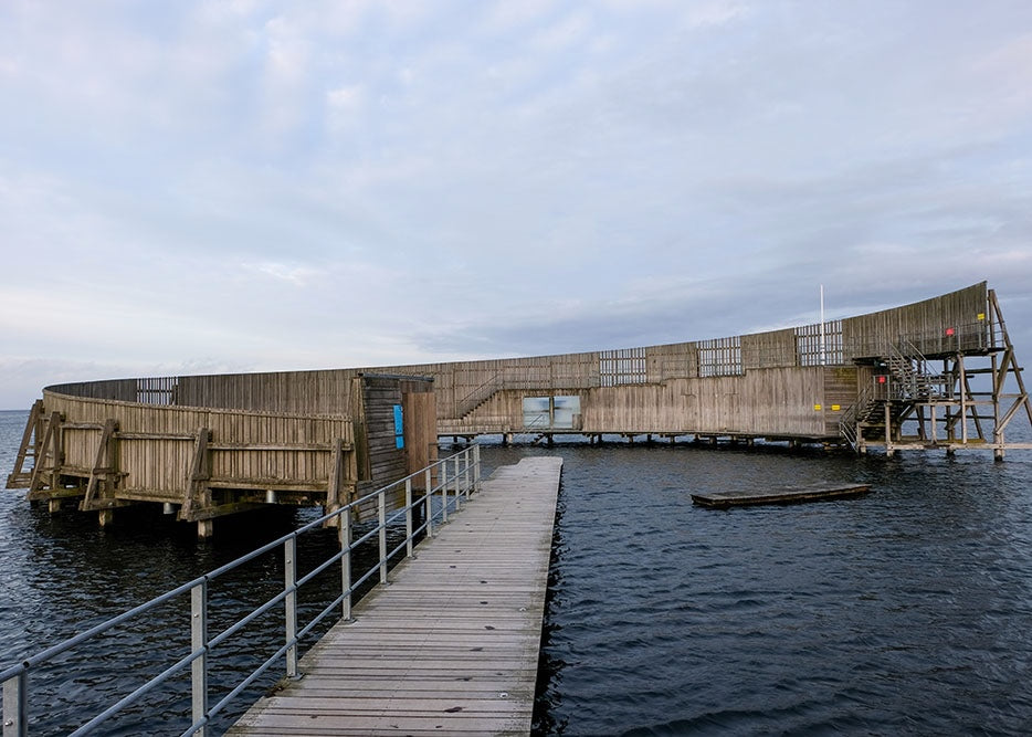 En trægang med rækværk fører til Sneglen Amager, et unikt cirkulært havbassin af Posters, hævet over roligt vand og med synlige trin til højre - ideelt til en rejseplakat eller plakat.