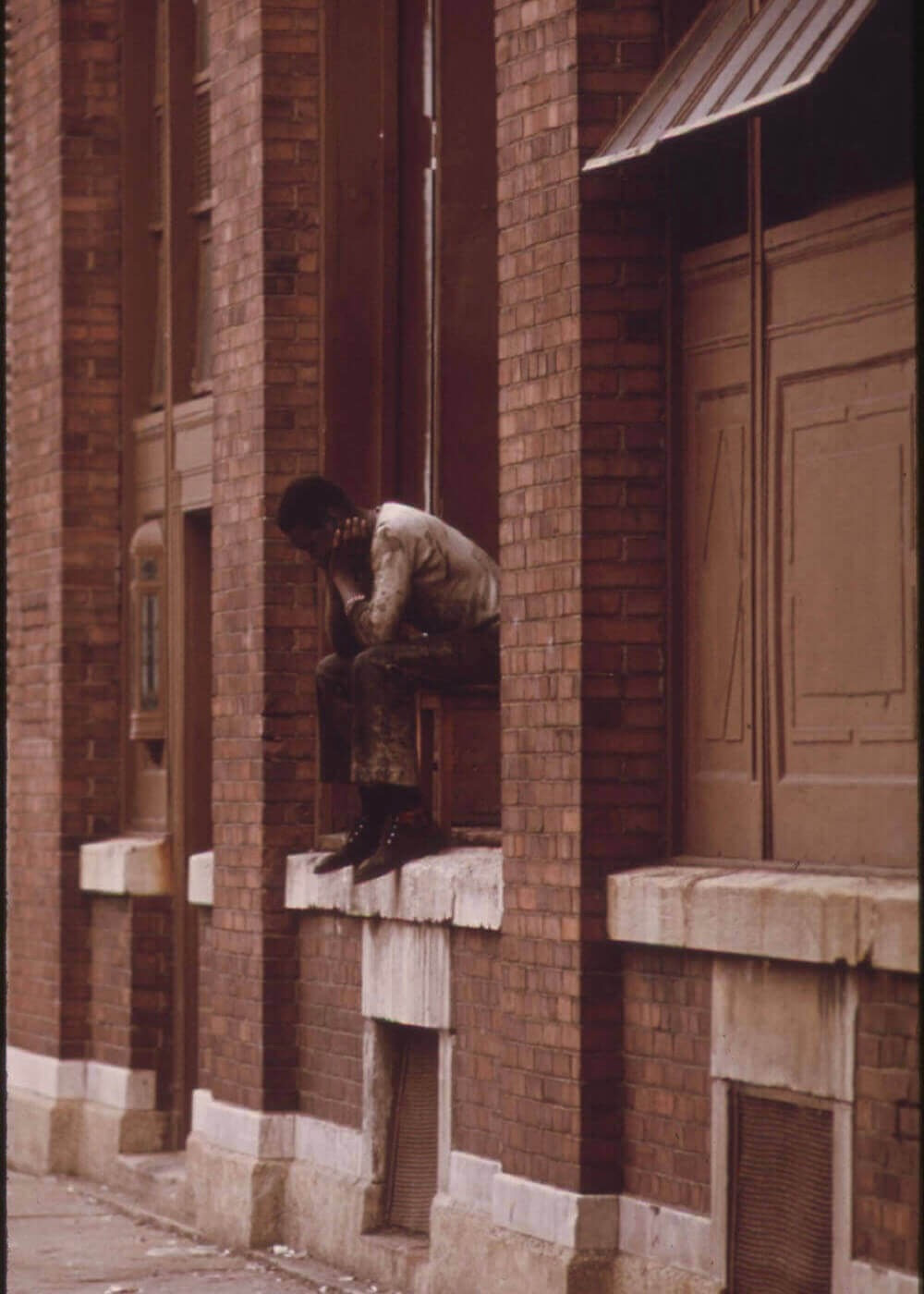 En mand sidder alene på en murstensbygnings vindueskarm, læner sig frem med hånden på hovedet, dybt optaget af tanker ved siden af en falmet plakat. Den tomme gade og bygning fremkalder den stille stemning i Posters' Tænkeren.