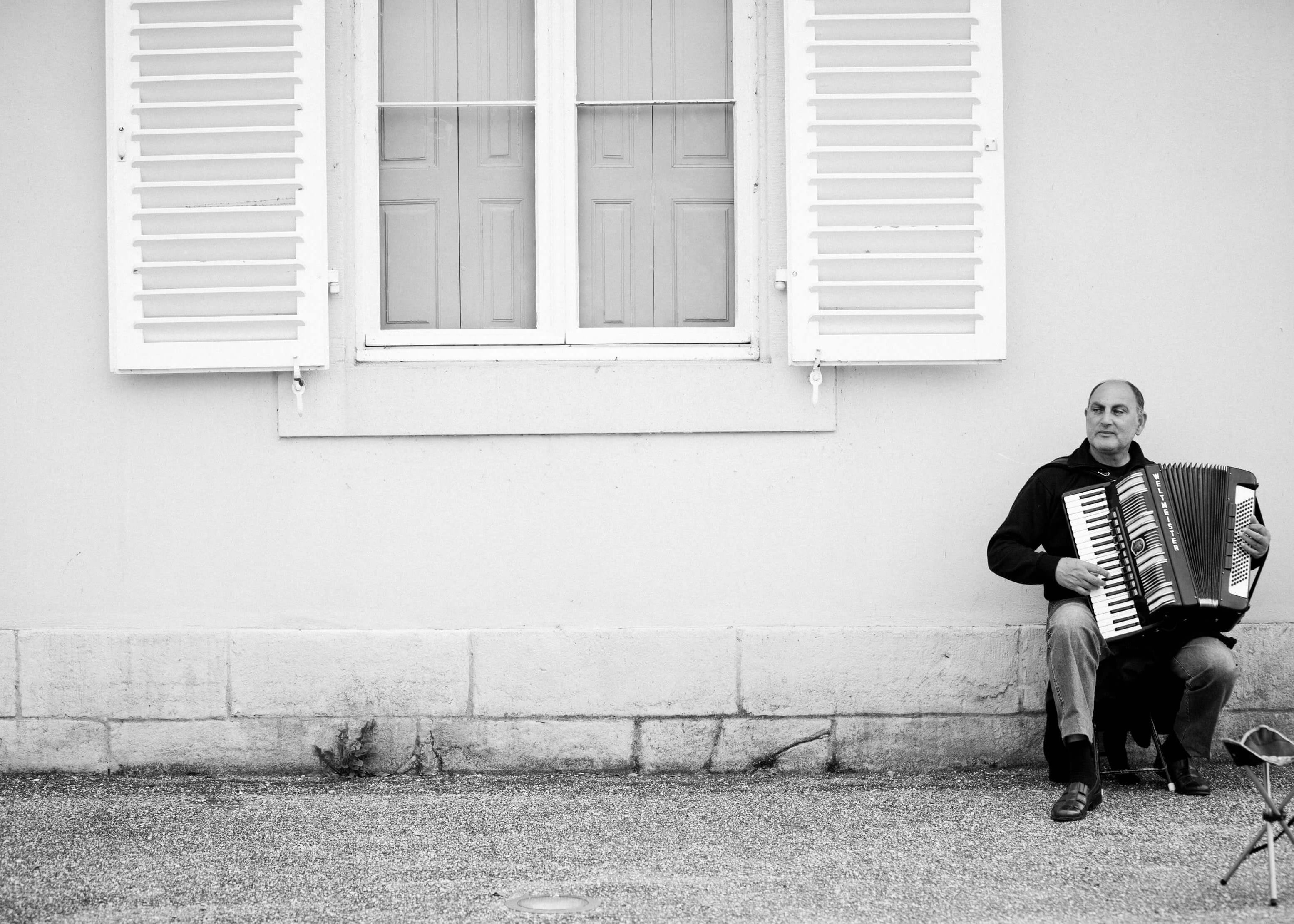 The Music Man" af Posters viser en mand på en skammel, der spiller harmonika ved siden af et åbent vindue på en almindelig væg; det sort-hvide design i plakatstil fremhæver hans placering til højre og fremkalder minimalistisk charme.