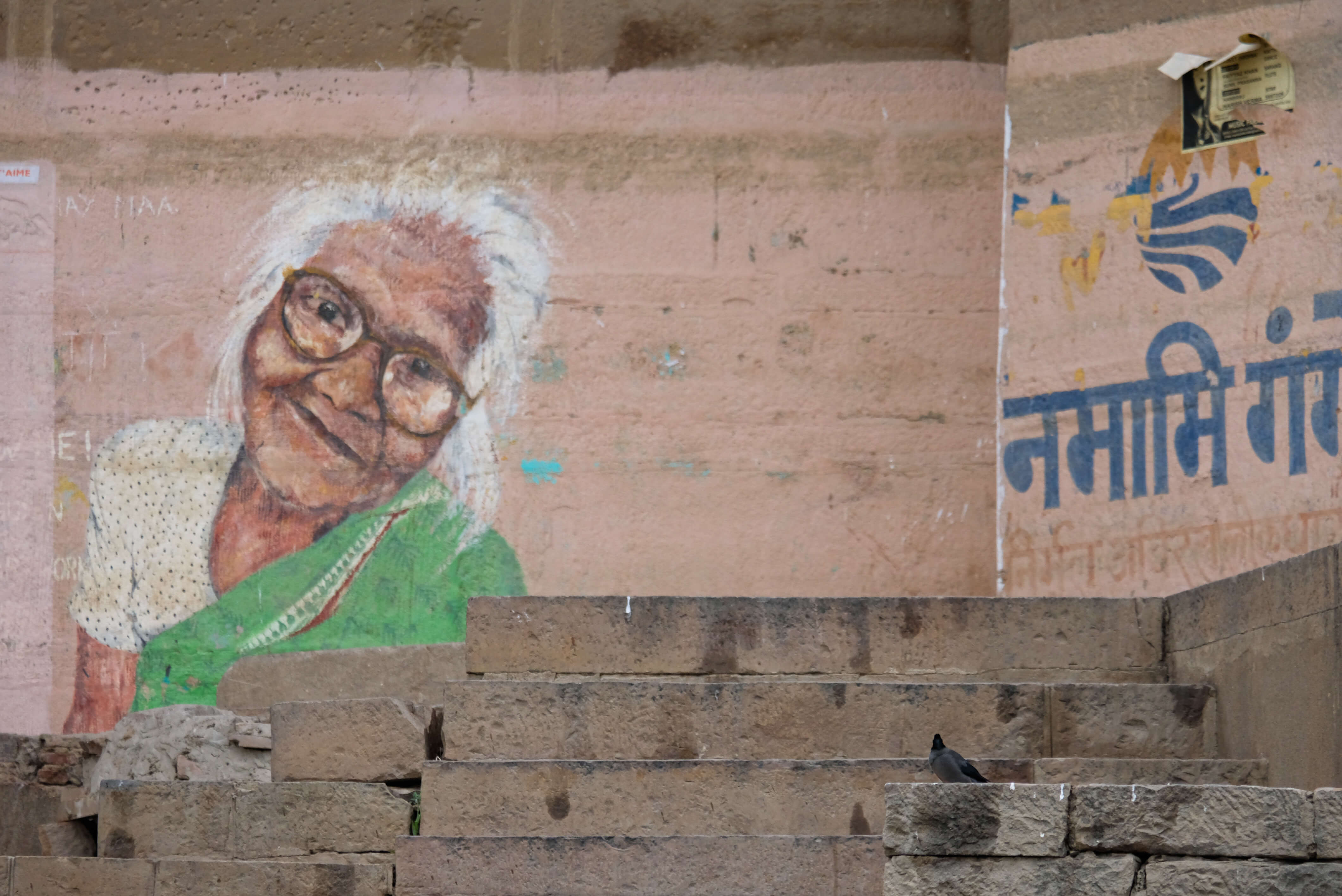 Wallart by Posters har et vægmaleri af en ældre kvinde i grøn sari og briller på en forvitret mur over stentrapper, med hindi-tekst, et håndmalet Plakat-logo og en due, der fuldender scenen.